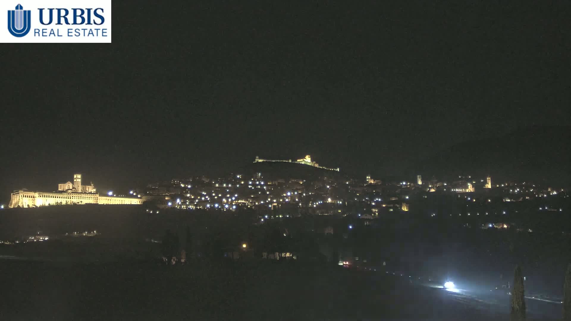 A nighttime view of a hilltop town illuminated by scattered lights, with a large, brightly lit building at the base of the hill.