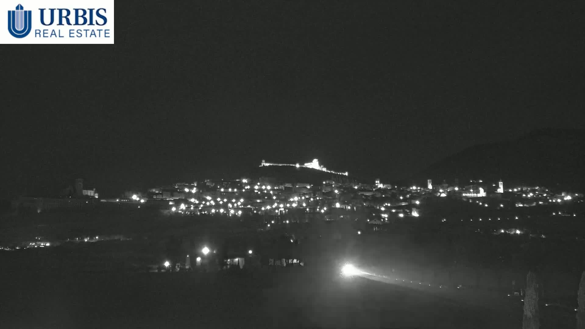 A nighttime, long shot shows a hilltop town illuminated by scattered lights, with a road and headlights visible in the foreground under a dark, clear sky.