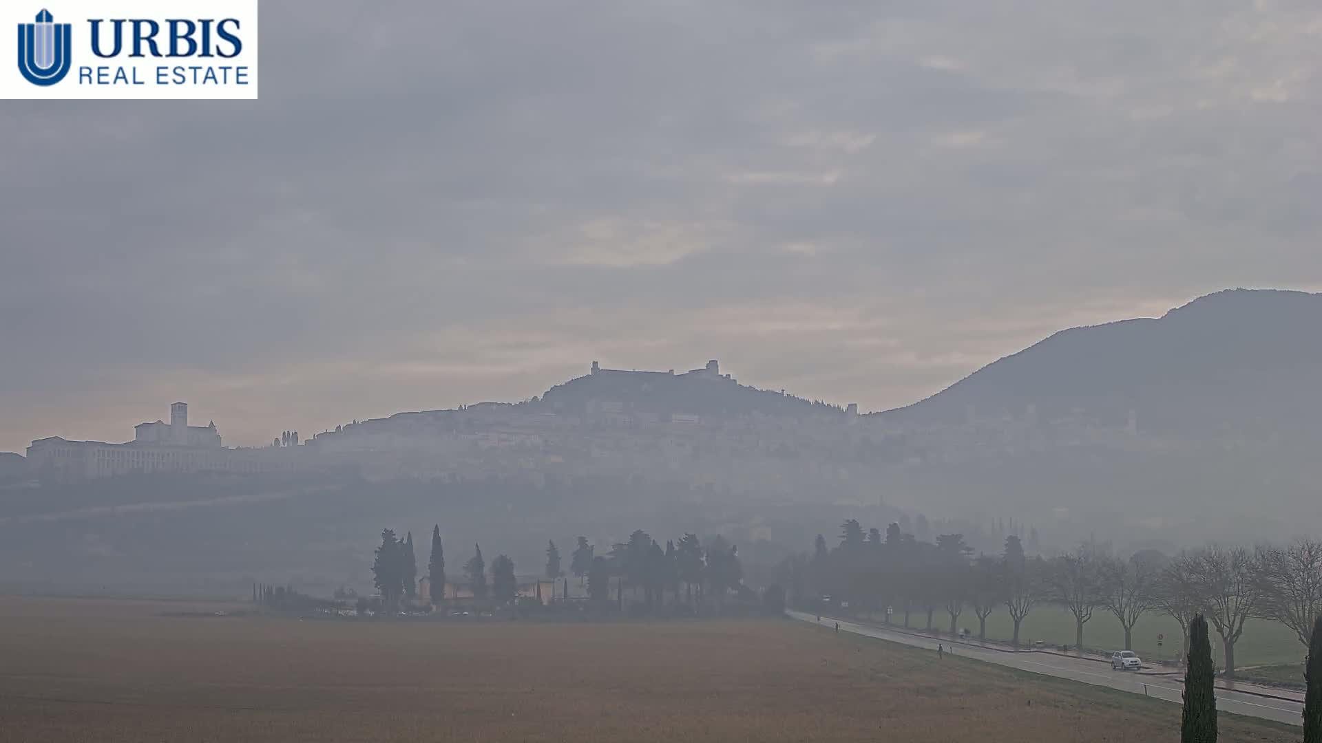 A hazy, overcast day reveals a distant hilltop town and a large, flat field in the foreground, with a road and sparse trees lining the bottom.