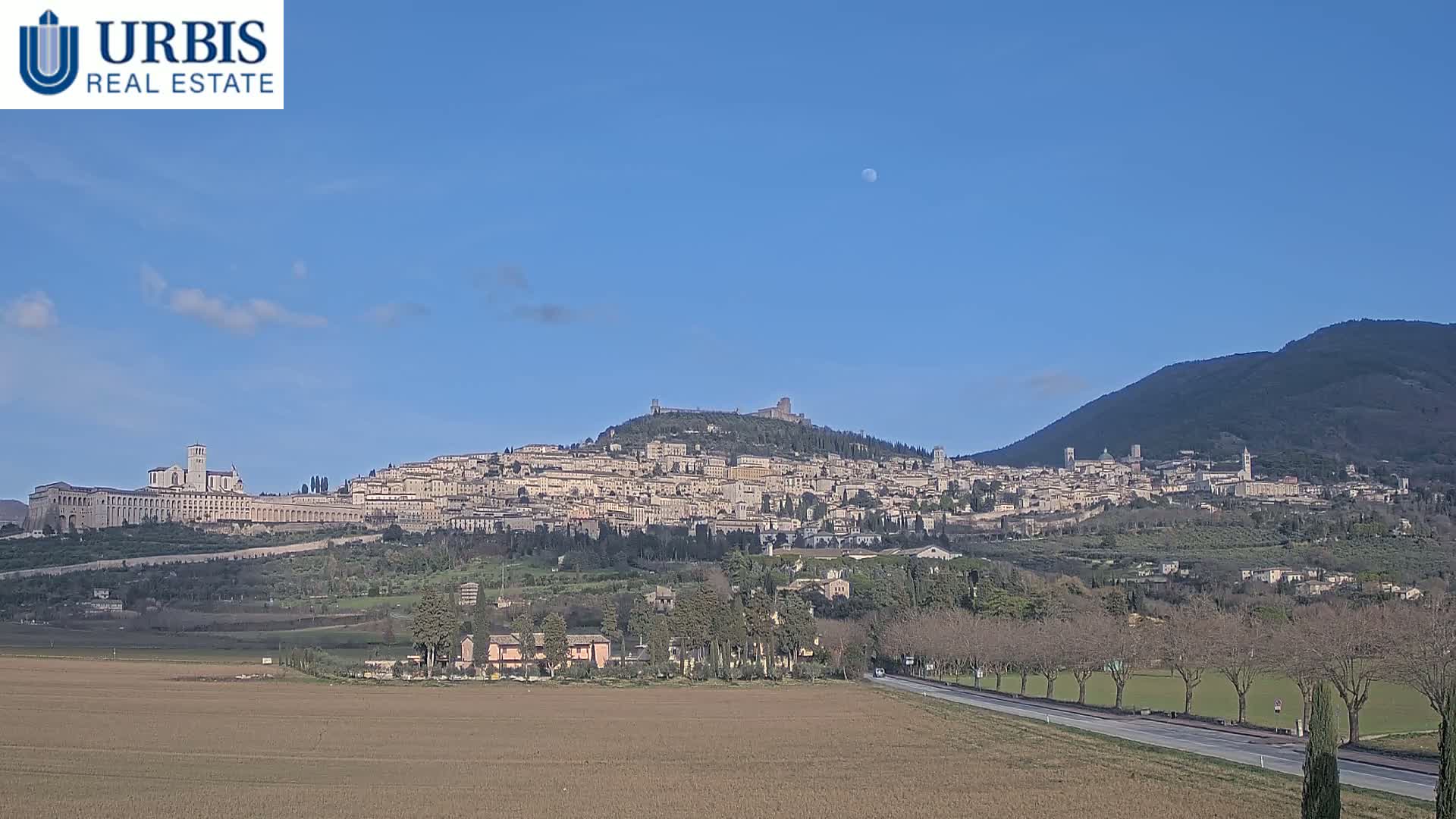 A hilltop town, nestled between mountains under a clear, sunny sky with a visible moon, is seen from a distance across a flat expanse of land with a road running through it.
