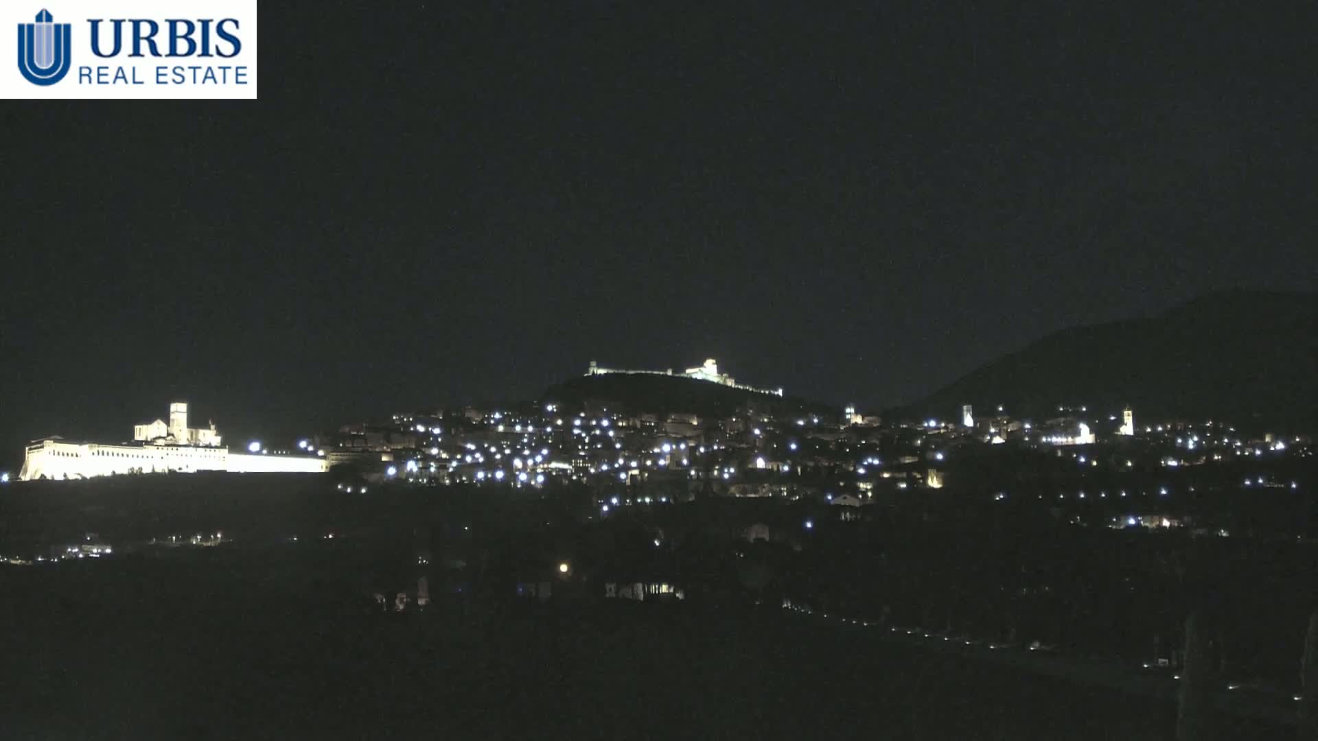 A nighttime view of a town nestled on a hill, illuminated by scattered lights, with a larger building complex brightly lit on a lower hill in the foreground under a clear, dark sky.