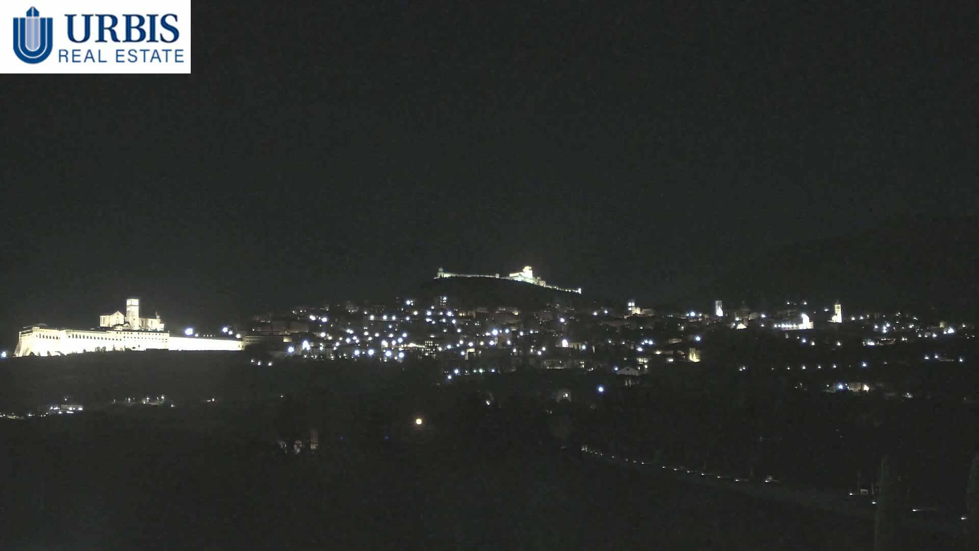 A nighttime view of a hilltop town with illuminated buildings, including a large, brightly lit structure on a lower hill.