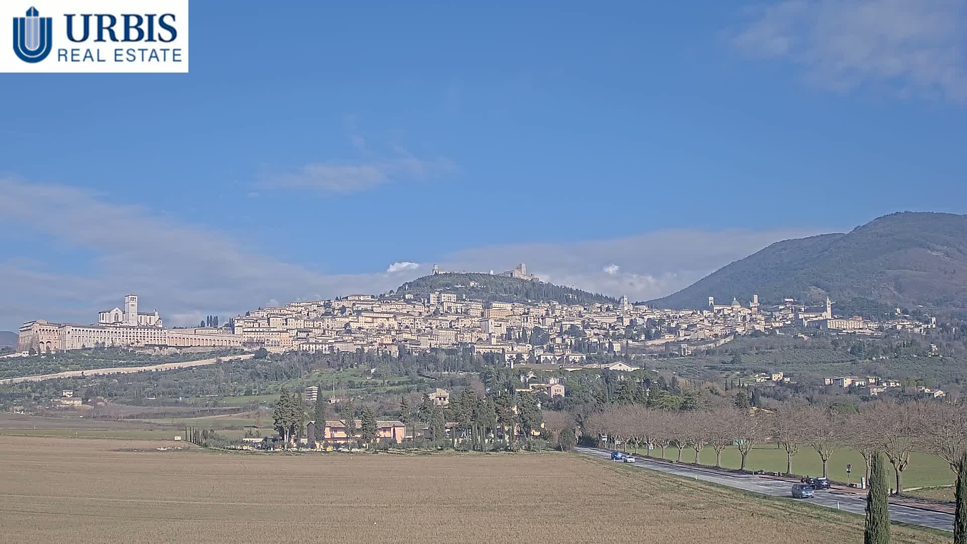 A hilltop town, with many buildings and several prominent structures, sits under a clear blue sky, with a mountain visible in the background and a flat, brown field in the foreground.