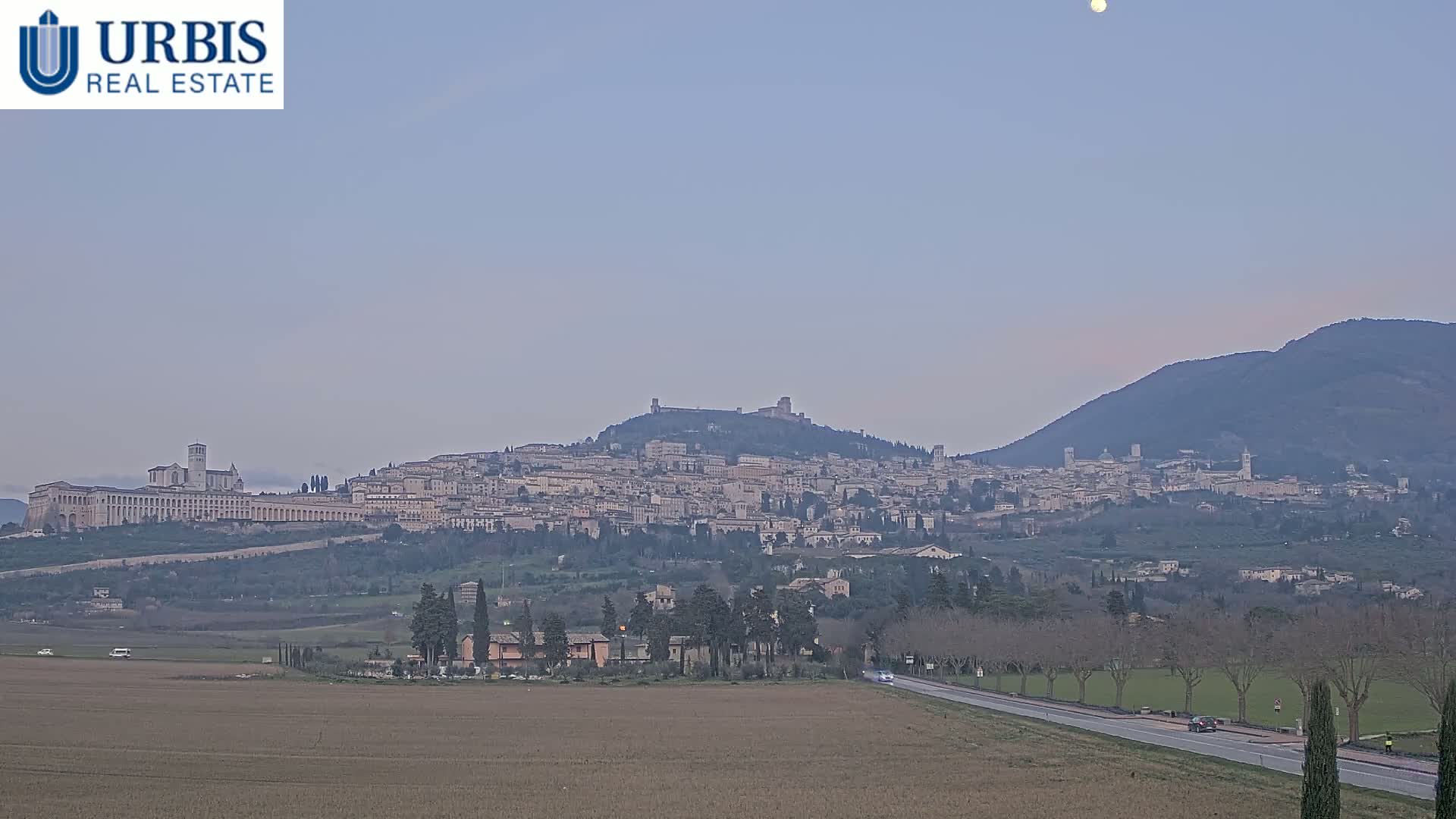 A hilltop town, featuring a large basilica, is visible in the distance under a clear, pale sky with a visible moon.