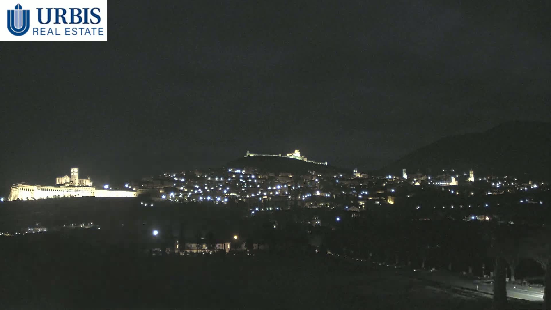 A nighttime view of a hilltop town illuminated by scattered lights, with a large, brightly lit building on a lower hill in the distance under a dark sky.