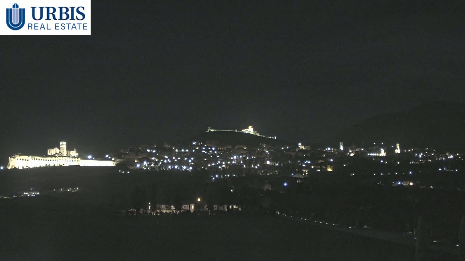 A nighttime view of a hilltop town illuminated by many small lights, with a large, brightly lit building on a lower hill in the foreground.