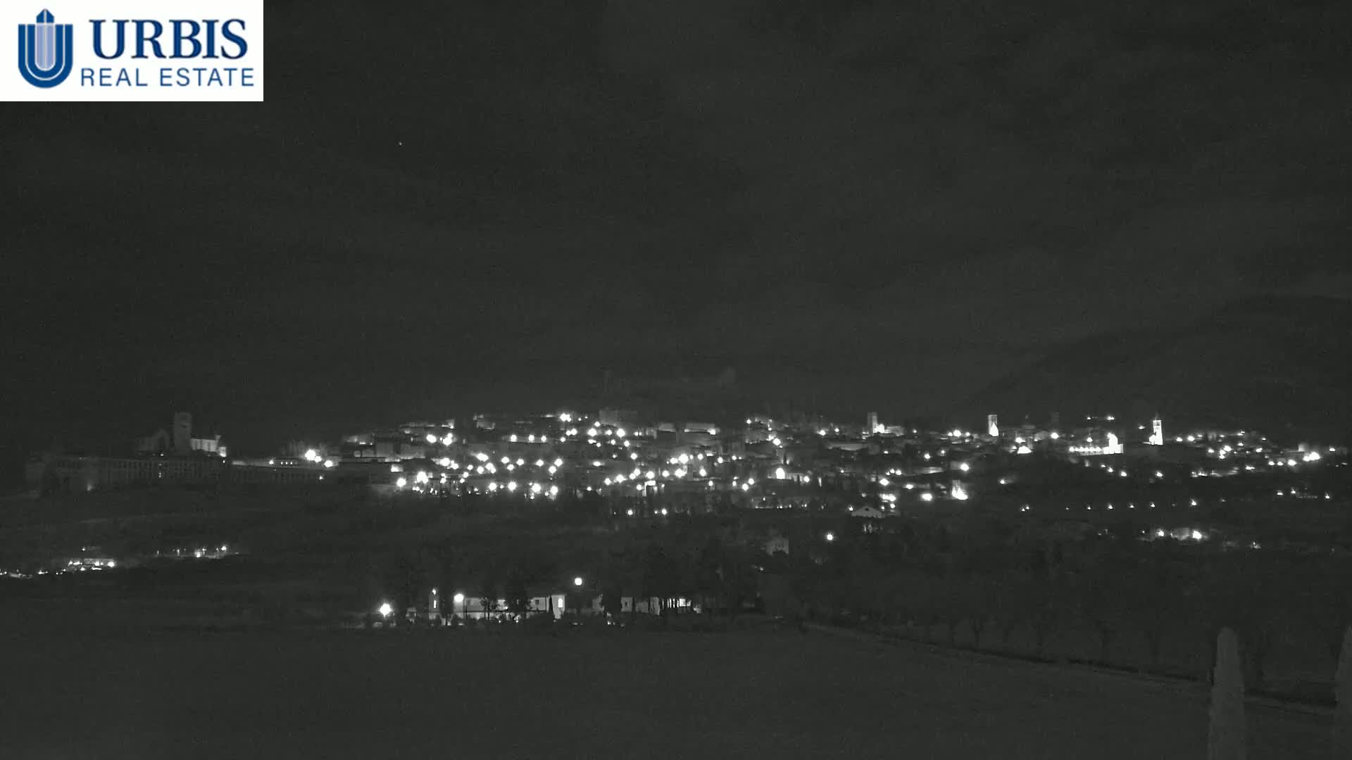 A nighttime long shot shows a town illuminated by scattered lights, nestled in a hilly landscape under a dark, mostly clear sky.