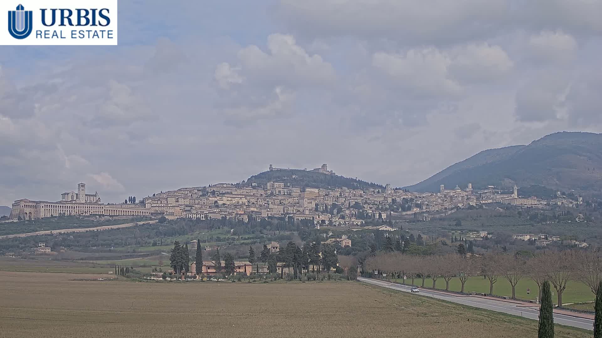 A sprawling hilltop town, under a cloudy sky, is viewed from a distance across a flat field with a road running through it.