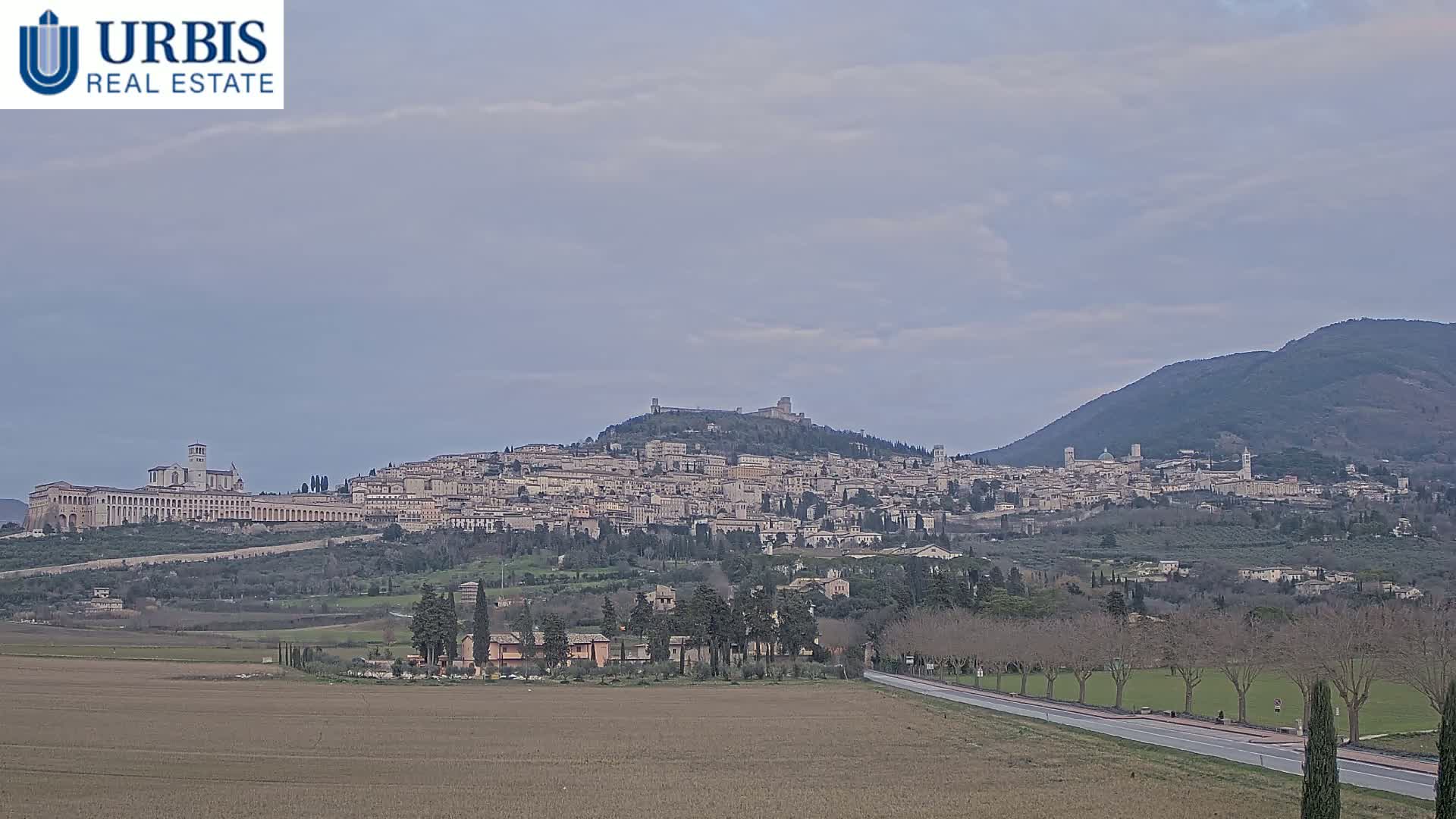 A sprawling hilltop town, under an overcast sky, is seen from a distance across a flat field and road.