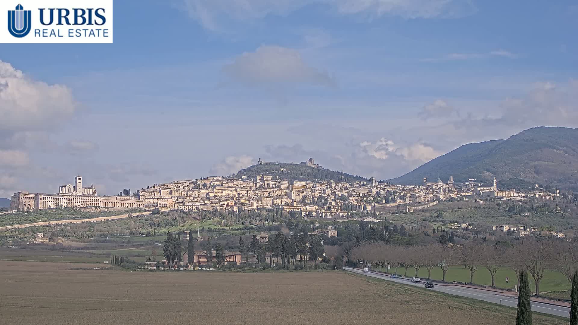 A large hilltop town is visible in the distance under a partly cloudy sky, with a flat, brown field and a road in the foreground.