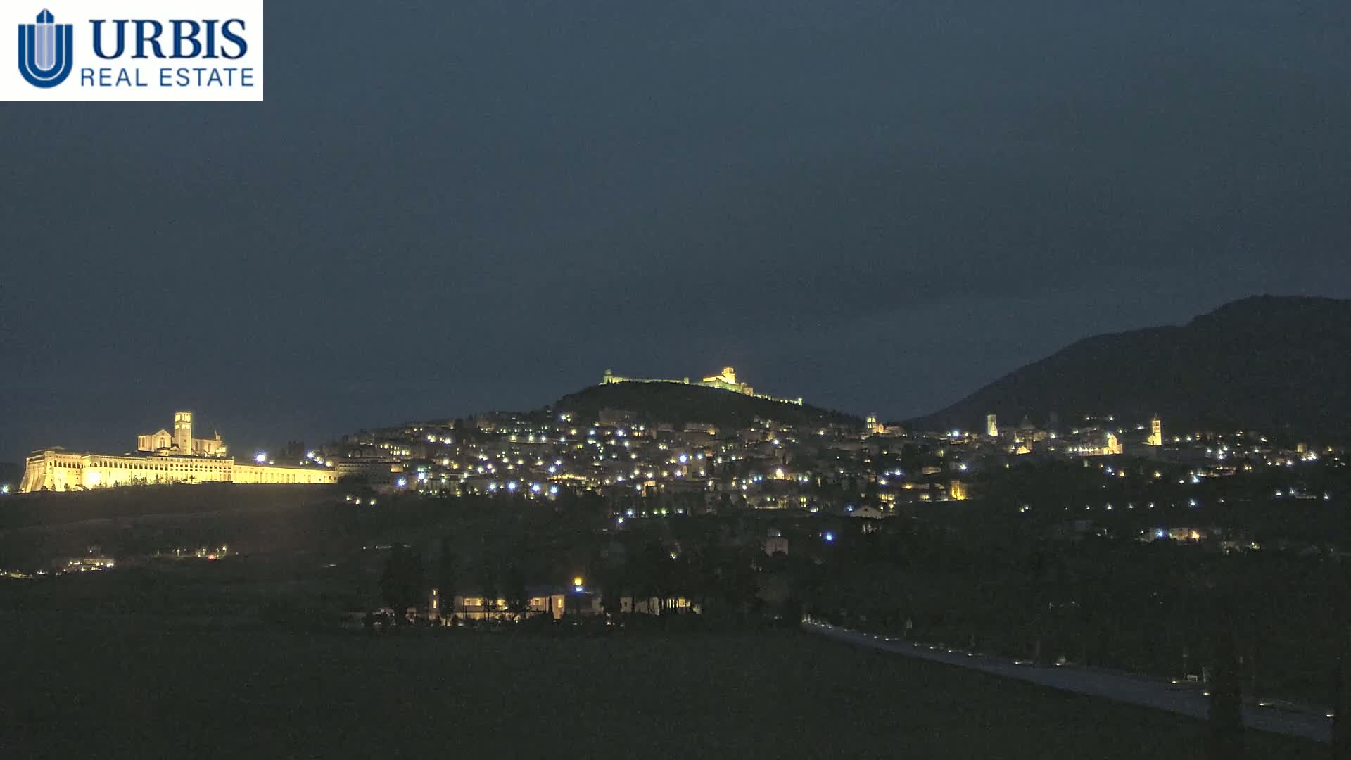 A hilltop town is illuminated at night, with a large, brightly lit building at the base of the hill, under a dark, clear sky.