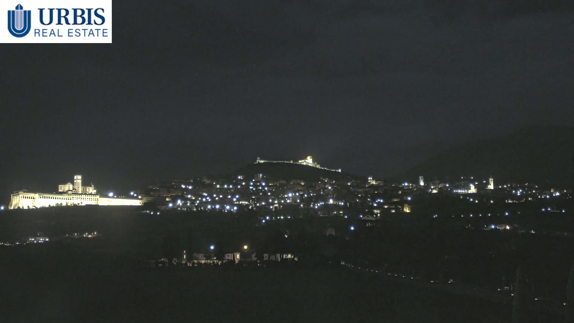 A nighttime view shows a hilltop town illuminated by scattered lights, with a larger, brighter structure situated on a lower hill in the foreground.