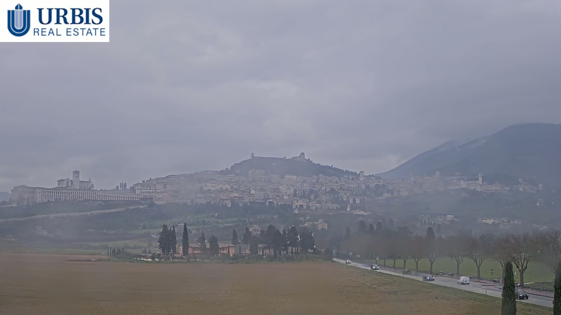 A hazy, overcast view shows a town nestled on a hill, with a large basilica visible, and a road with cars in the foreground.