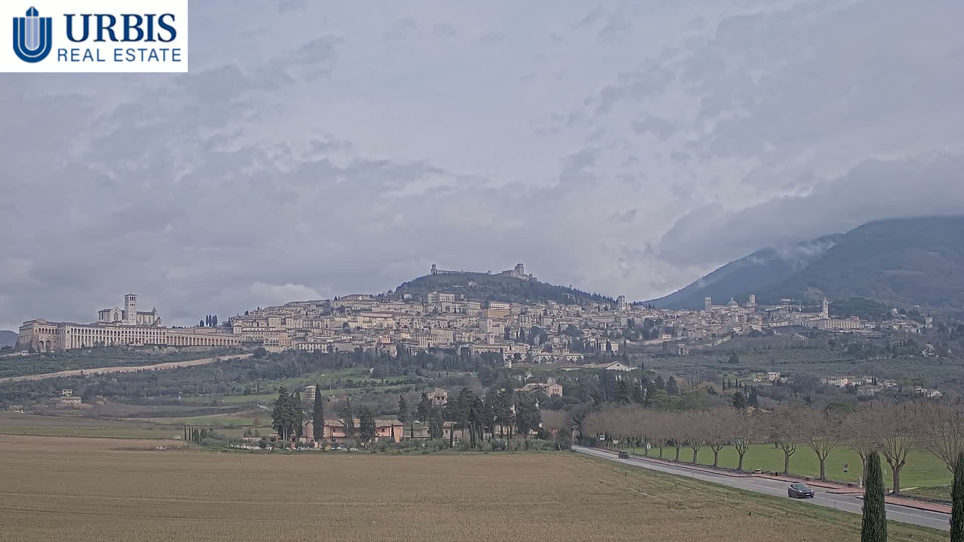 A large hilltop town, under a mostly cloudy sky, is viewed from a distance across a flat field and a road.