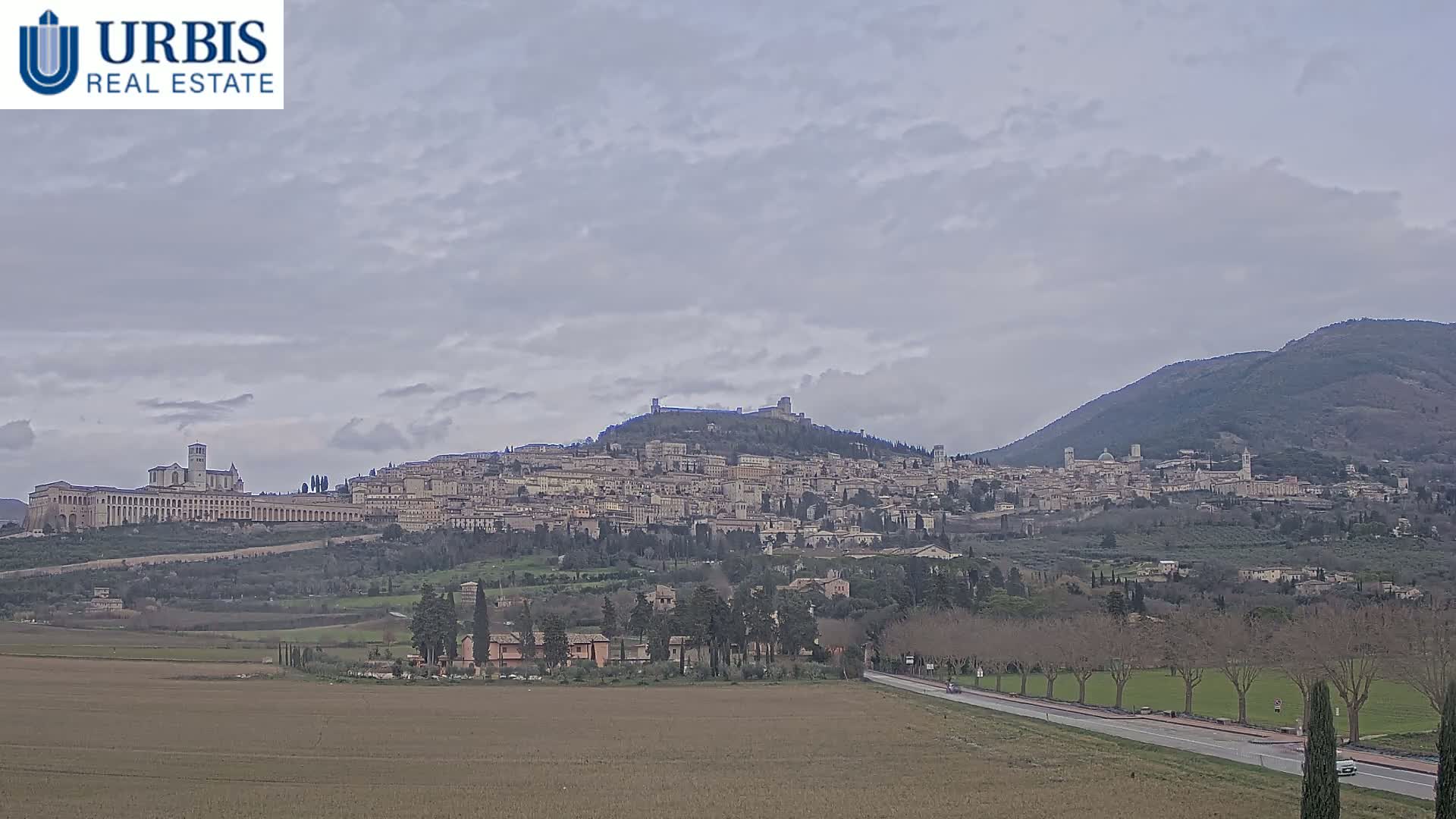 A hilltop town, nestled between rolling hills under a cloudy sky, is viewed from a distance across a flat field.