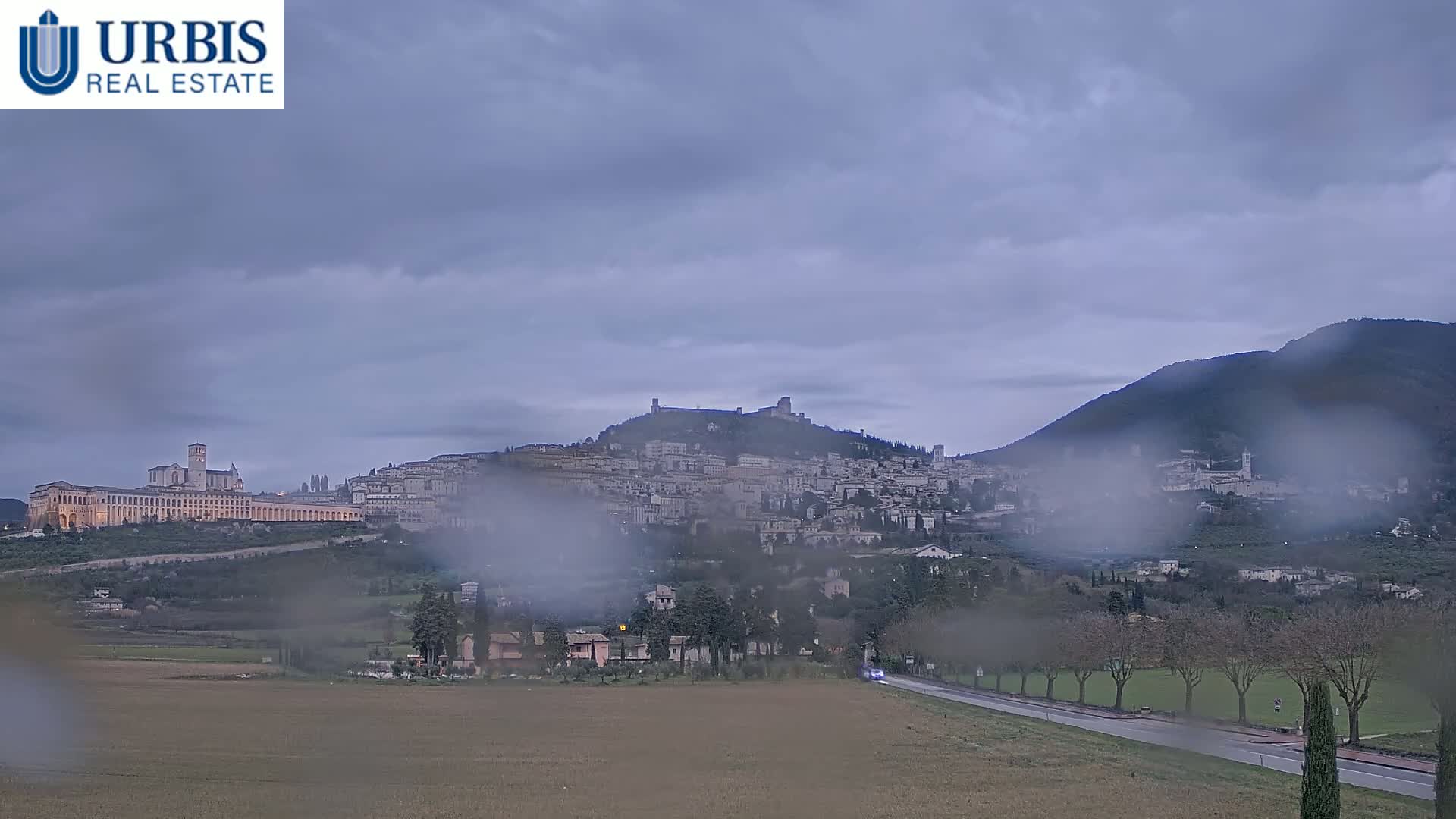 A hilltop town with a large basilica is visible through rain-streaked glass, under a cloudy sky.