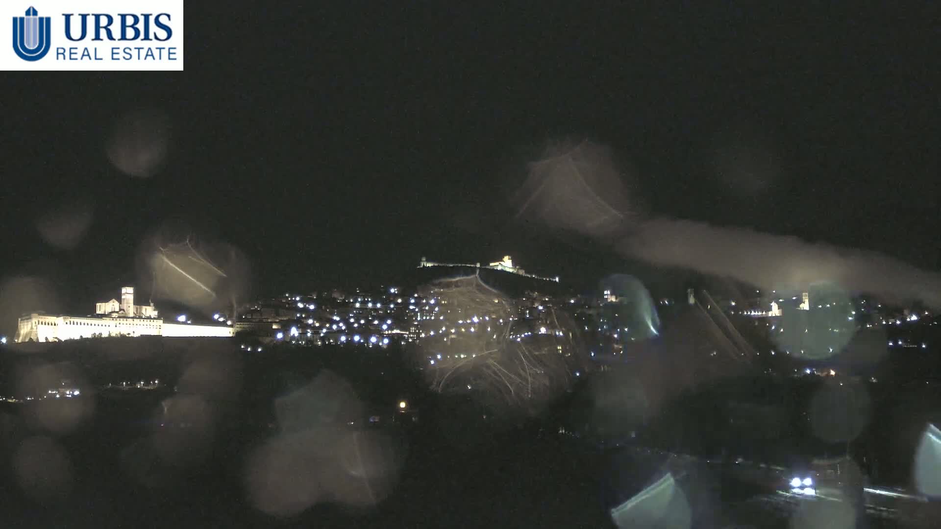 A nighttime view of a city on a hill, illuminated by lights, seen through rain-covered glass.