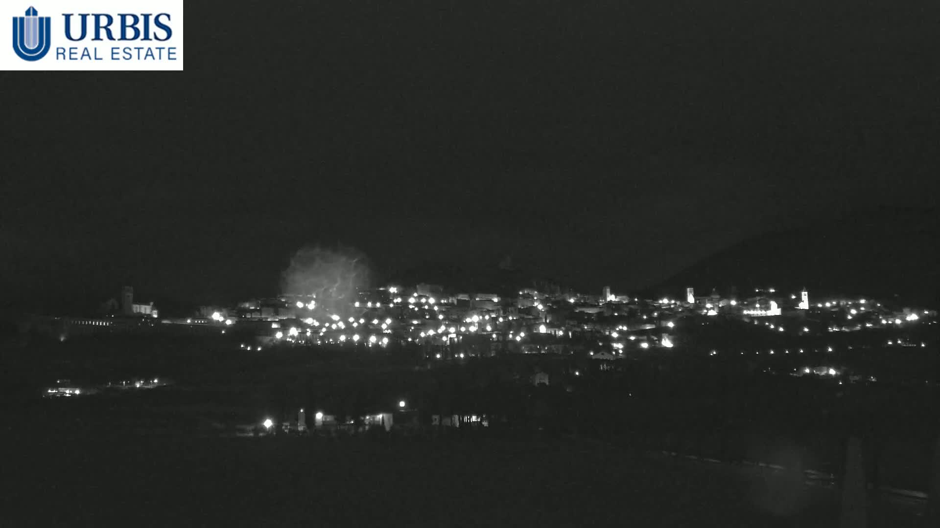 A nighttime long shot of a town nestled in a dark hill, illuminated by scattered lights under a cloudy sky.