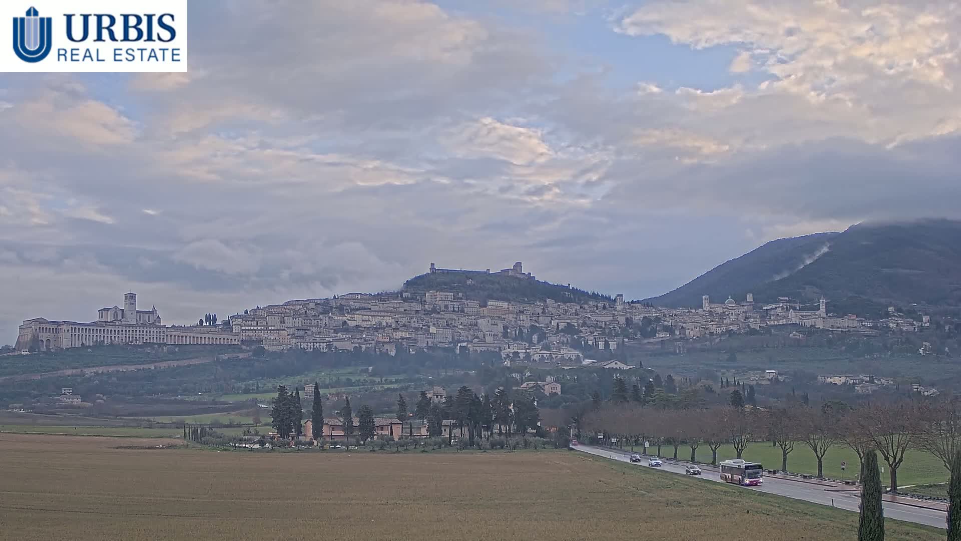 A large hilltop town, partially obscured by haze, is visible under a cloudy sky, with a flat field and road in the foreground.