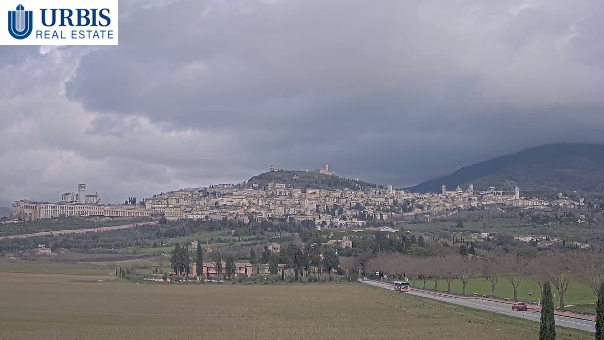 A hilltop town with numerous buildings, under a cloudy sky, is viewed from a distance across a flat, brown field and a road with sparse traffic.