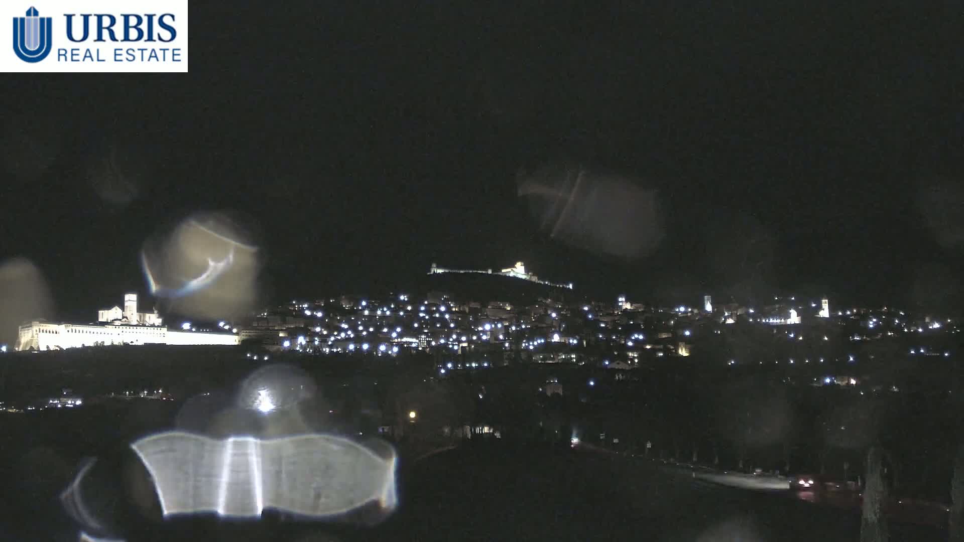 A nighttime view of a town nestled in a valley, illuminated by scattered lights, with a large building on a hill in the background, all seen through rain-covered glass.