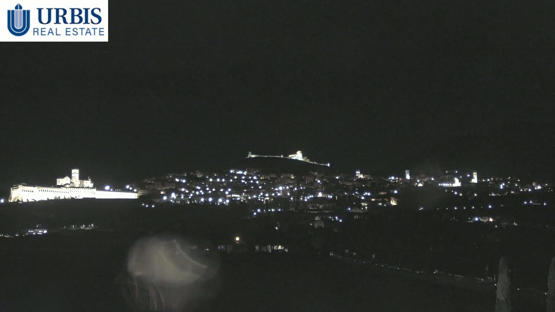 A nighttime long shot of a hilltop town illuminated by numerous lights, with a large, brightly lit structure at the base of the hill.
