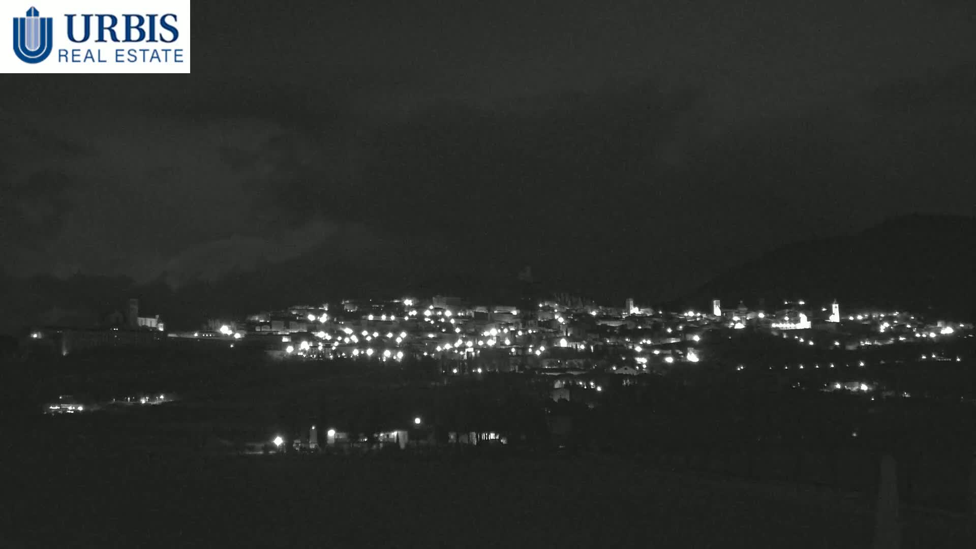 A nighttime, long-distance view of a town nestled in a valley under a dark, cloudy sky, illuminated by numerous small lights.