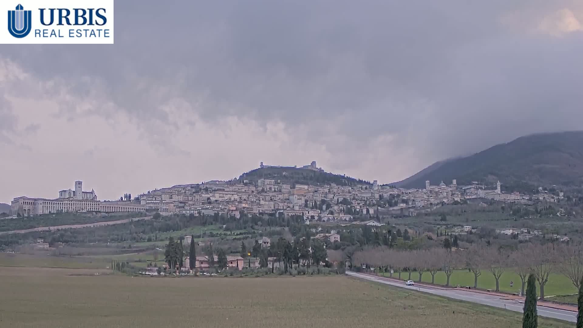 A hilltop town with numerous buildings, including a large basilica, is visible under an overcast sky, with flat farmland in the foreground.