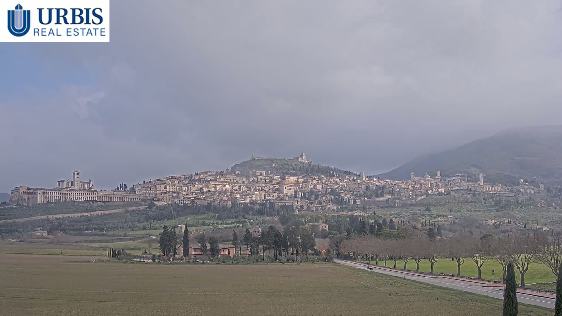 A sprawling town nestled on a hill, under an overcast sky, is viewed from a distance across a flat, sparsely vegetated plain.
