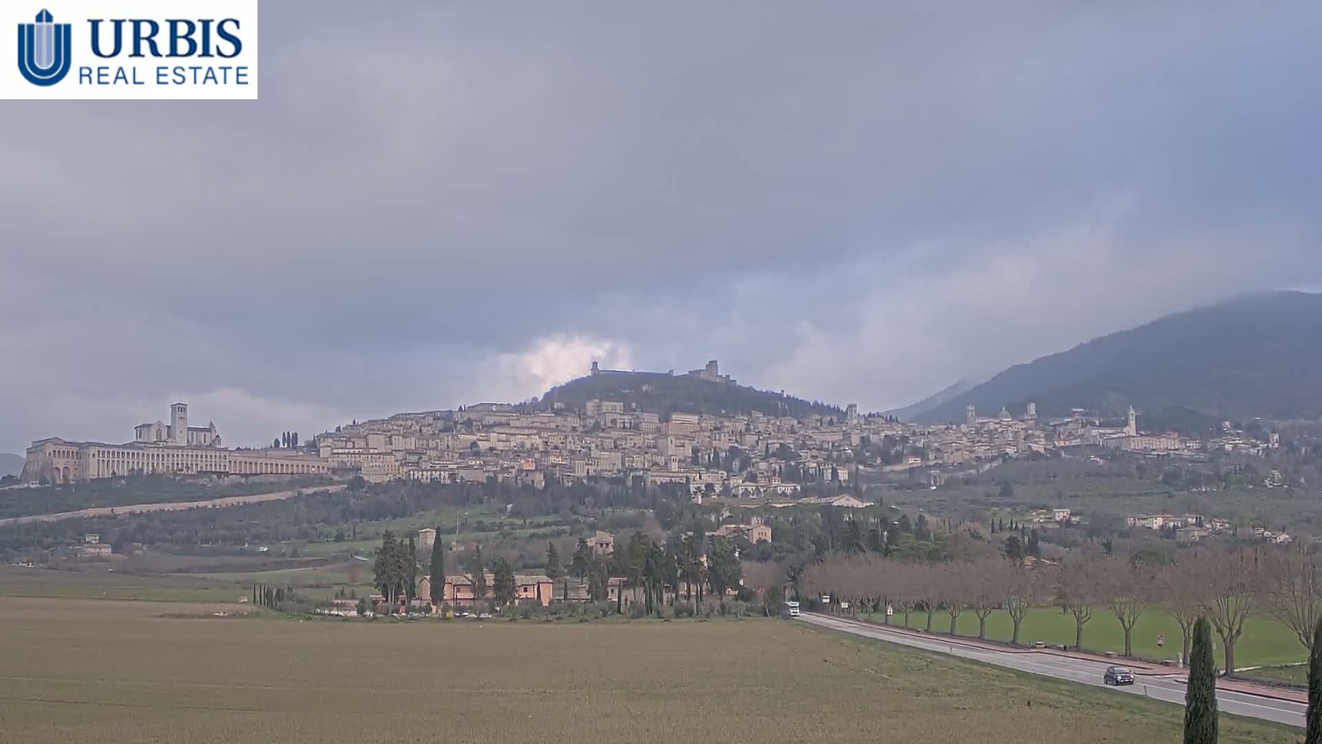 A large town sits atop a hill under a cloudy sky, with a flat, brown field and a road in the foreground.