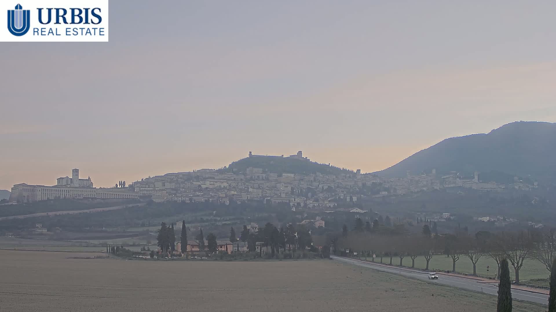 A hazy, pale sunrise illuminates a hilltop town nestled between two mountains, with a flat, frost-covered field in the foreground.