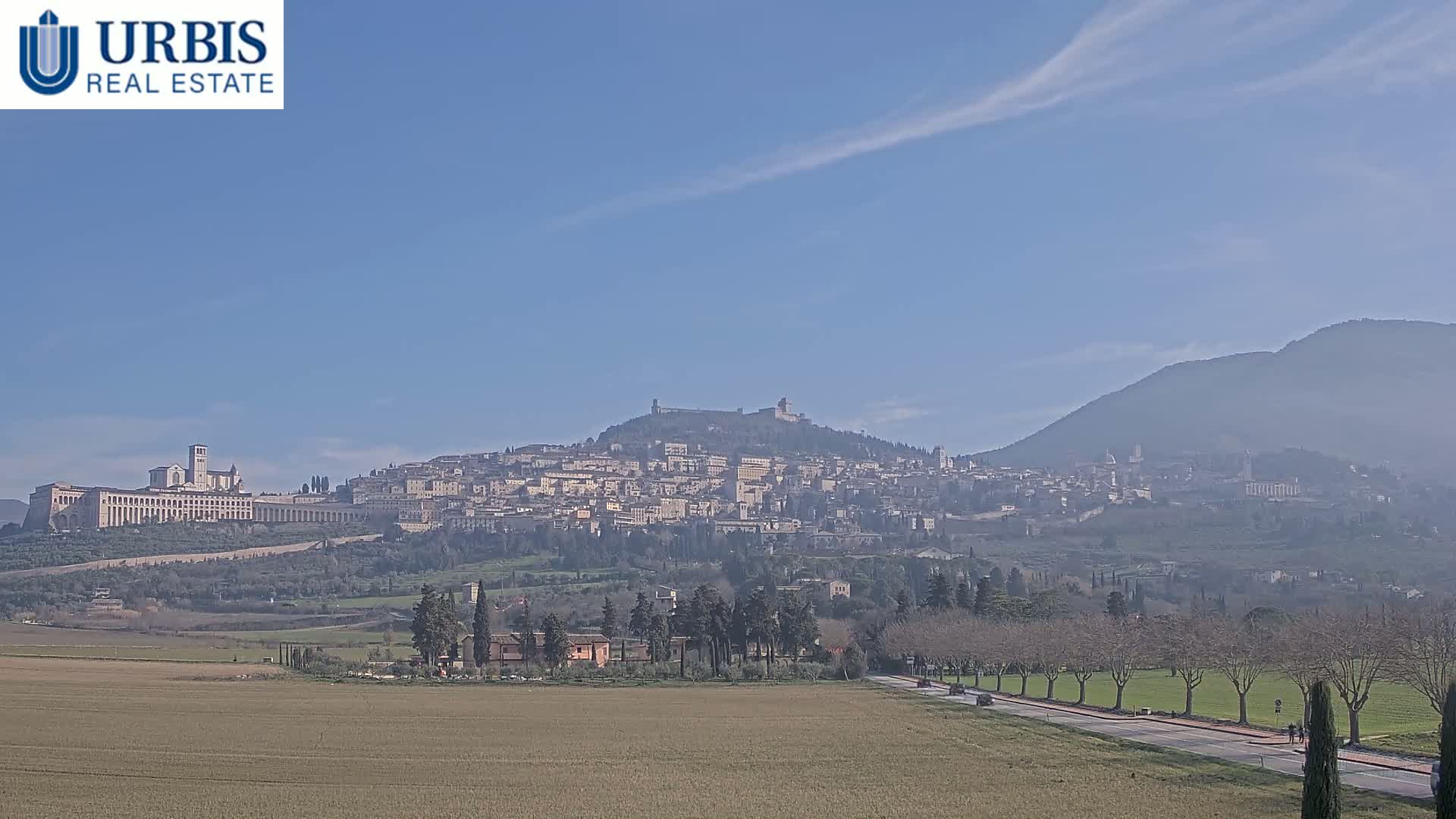 A hilltop town, possibly Assisi, is visible in the distance under a clear blue sky, with a flat, brown field in the foreground.
