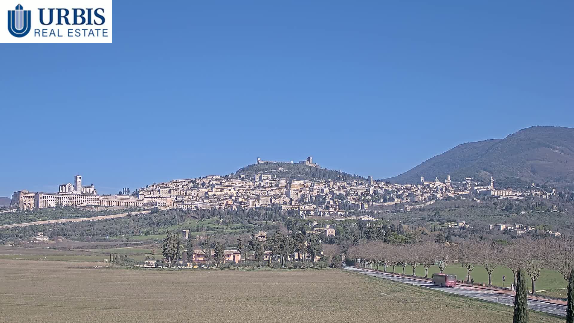 A large town sits atop a hill under a clear blue sky, with a flat, brown field and a road in the foreground.