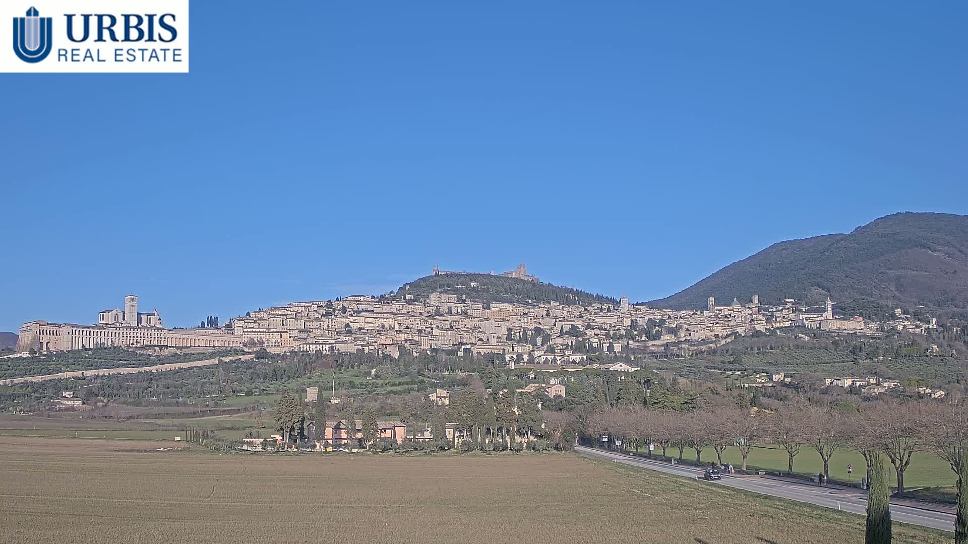 A hilltop town with numerous buildings, including a large basilica, is visible under a clear blue sky, with a flat expanse of farmland and a road in the foreground.
