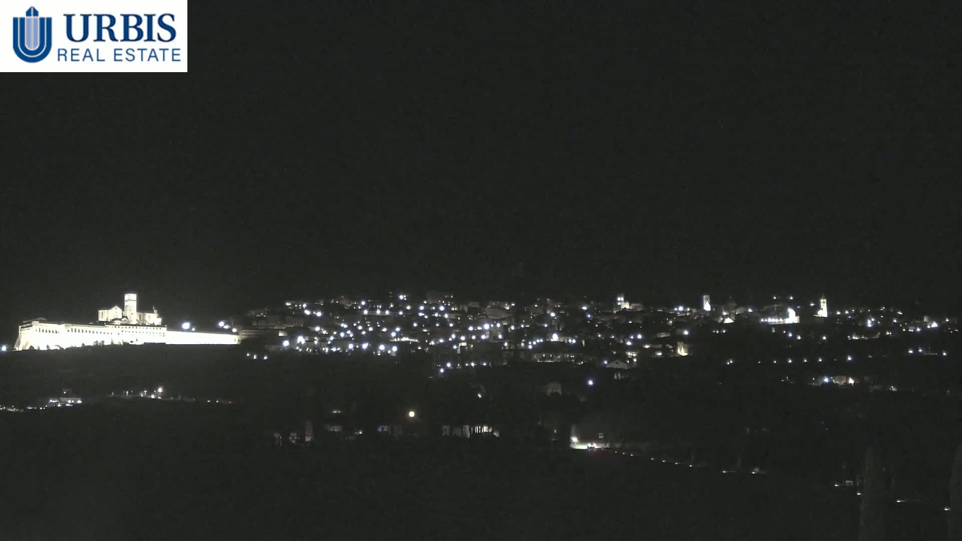 A brightly lit town and a large, illuminated building sit atop a hill at night under a clear, dark sky.
