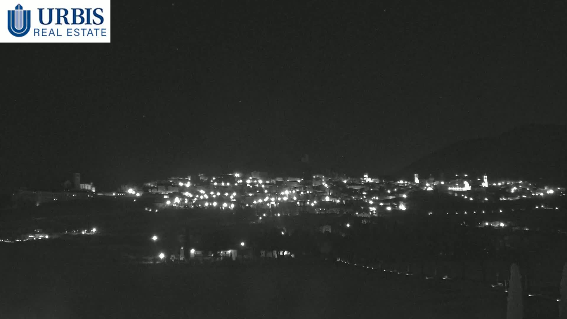 A nighttime long shot shows a small town nestled on a hill, its lights scattered across the landscape under a clear, dark sky.