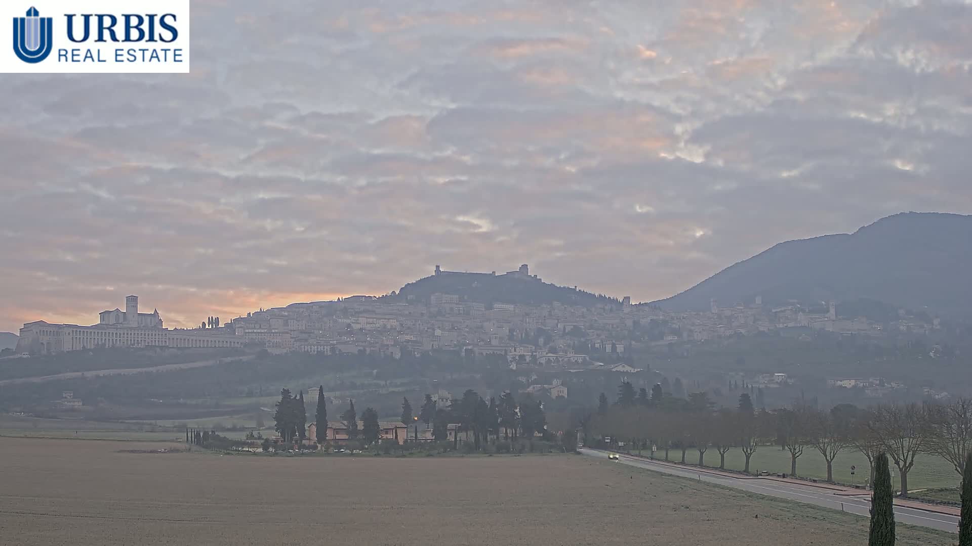 A hazy sunrise illuminates a hilltop town and a large, flat-topped mountain in the distance, under a mostly cloudy sky.