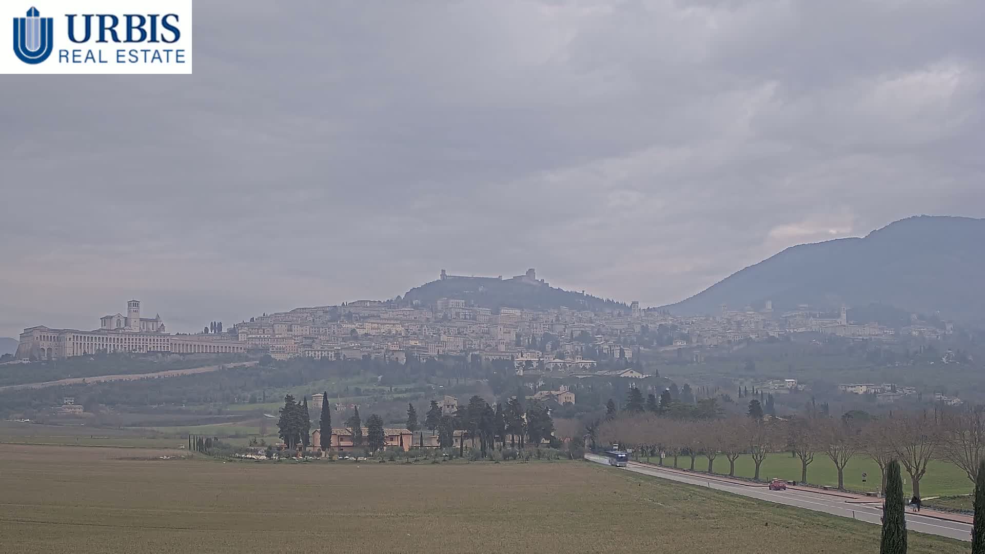A hazy, overcast day reveals a distant hilltop town with a large basilica, surrounded by a flat landscape with sparse trees and a road.