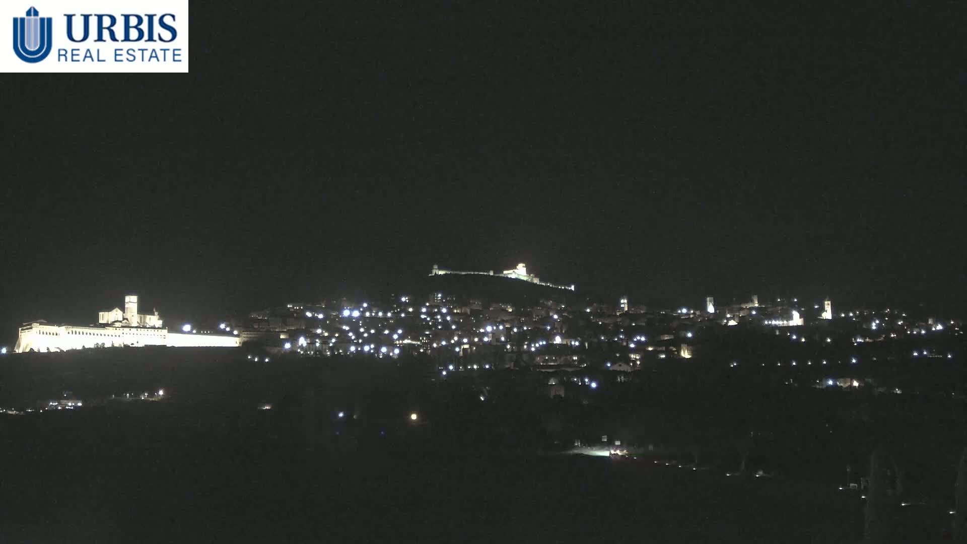 A nighttime view shows a hilltop town illuminated by scattered lights, with a large, brightly lit structure at the base of the hill.