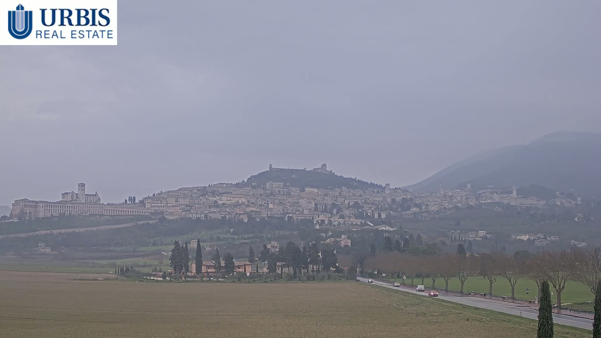 A hazy view of Assisi, Italy, sits on a hilltop, with a long, flat road and fields in the foreground.