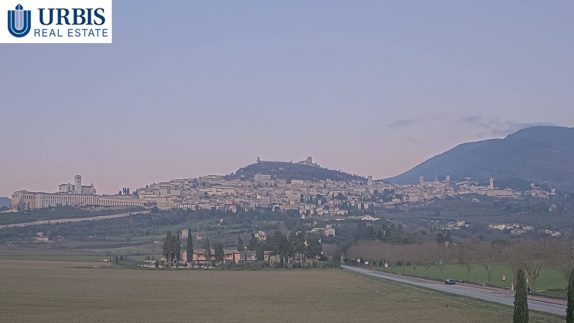 A large hilltop town is visible in the distance under a pale, dusky sky, with a flat field and road in the foreground.