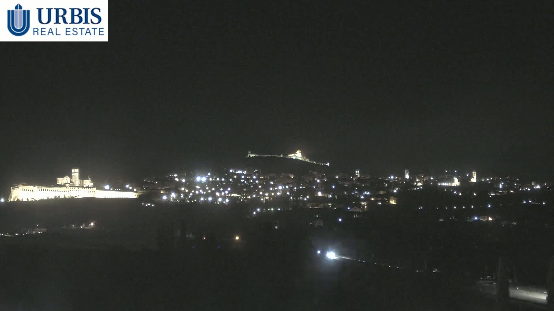 A nighttime view of a hilltop town, illuminated by scattered lights, with a large, brightly lit building at the base of the hill.