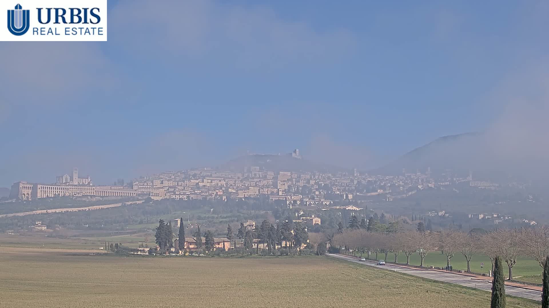 A hazy view of a distant city nestled in a valley, partially obscured by fog, with a flat, dry field in the foreground and a road leading towards the city.