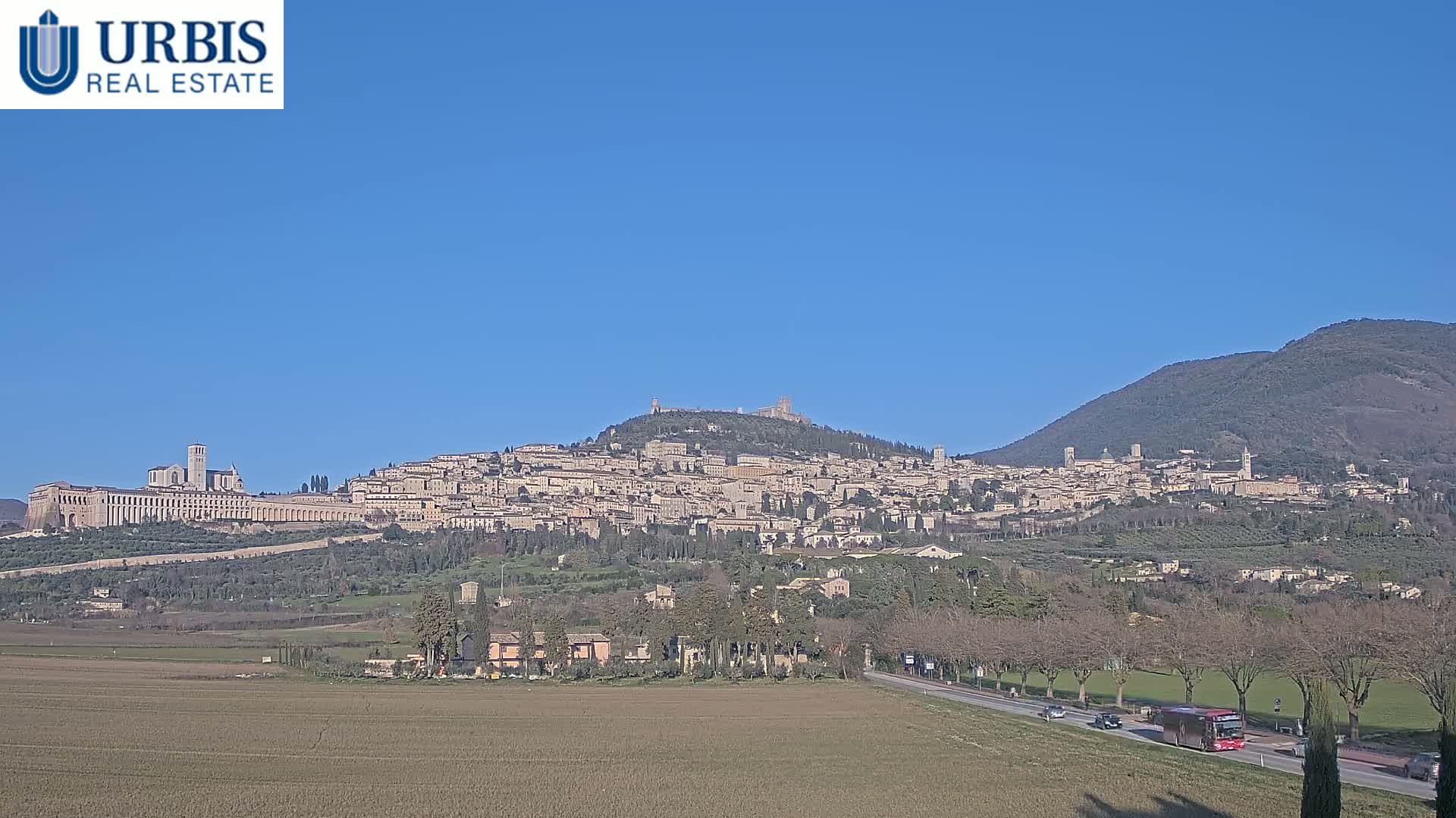 A large hilltop town, partially surrounded by mountains, is viewed from a distance on a clear, sunny day.