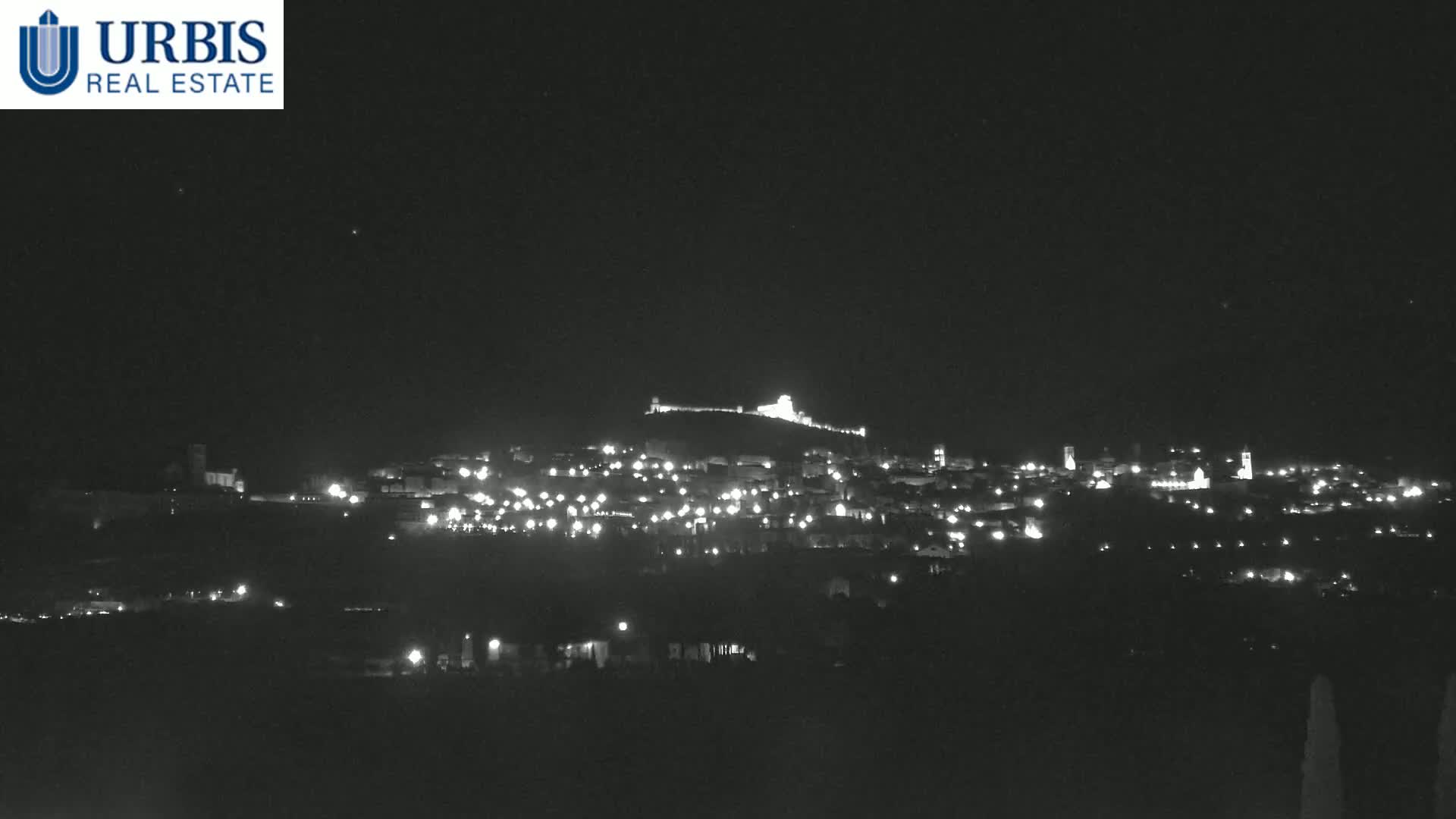 A nighttime long shot shows a hilltop town illuminated by many small lights, under a clear, dark sky.