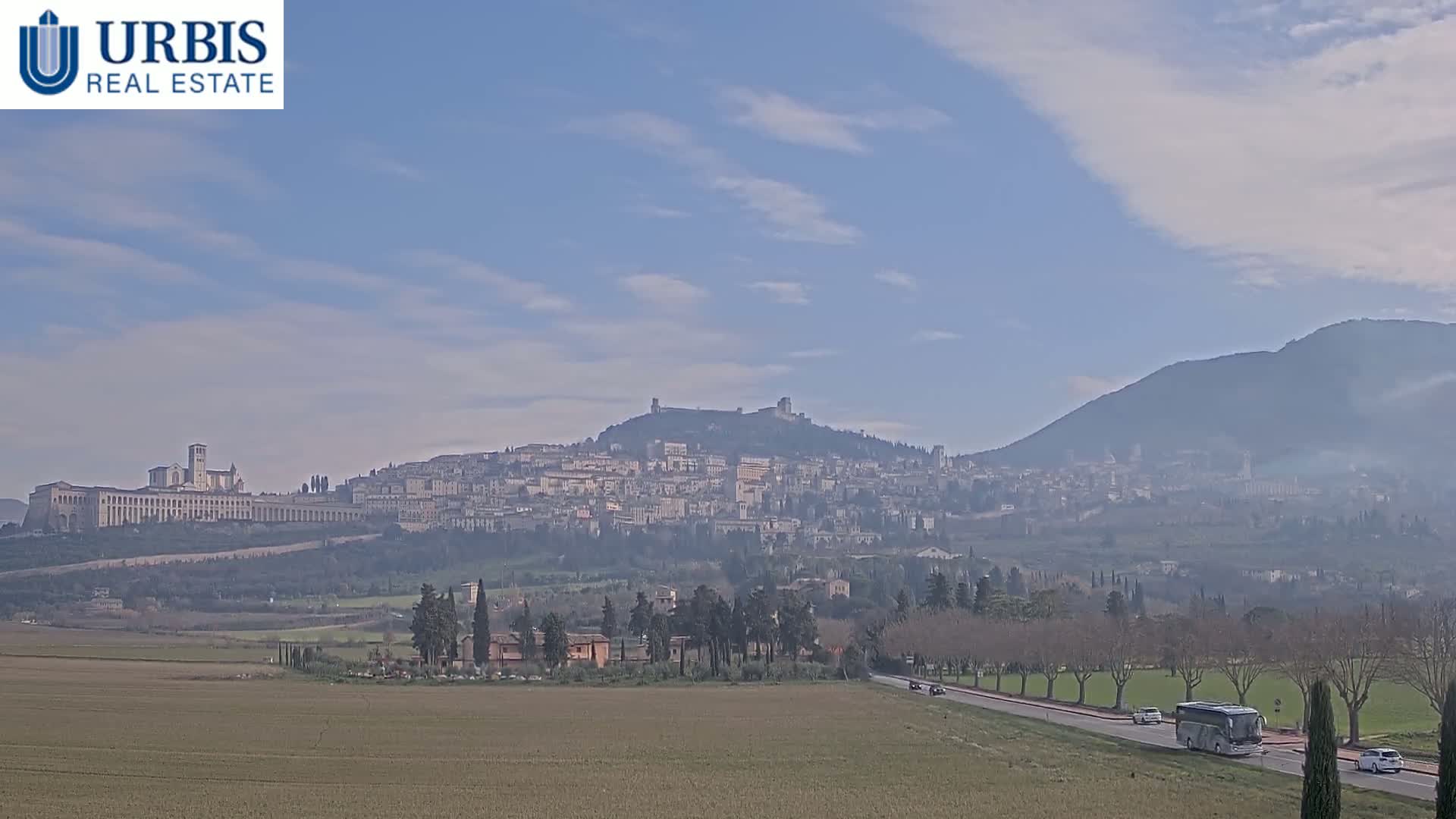 A hazy, sunny day reveals a sprawling hillside town with a large basilica and a mountain in the background, viewed from a flat, brown field with a road and sparse trees in the foreground.