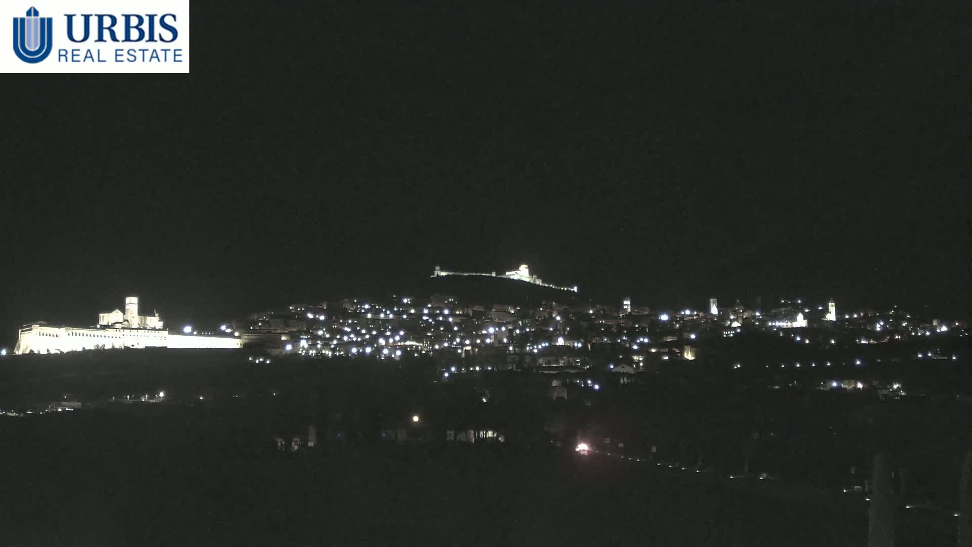 A nighttime view shows a hilltop town illuminated by lights, with a large, brightly lit building on a lower hill in the foreground.