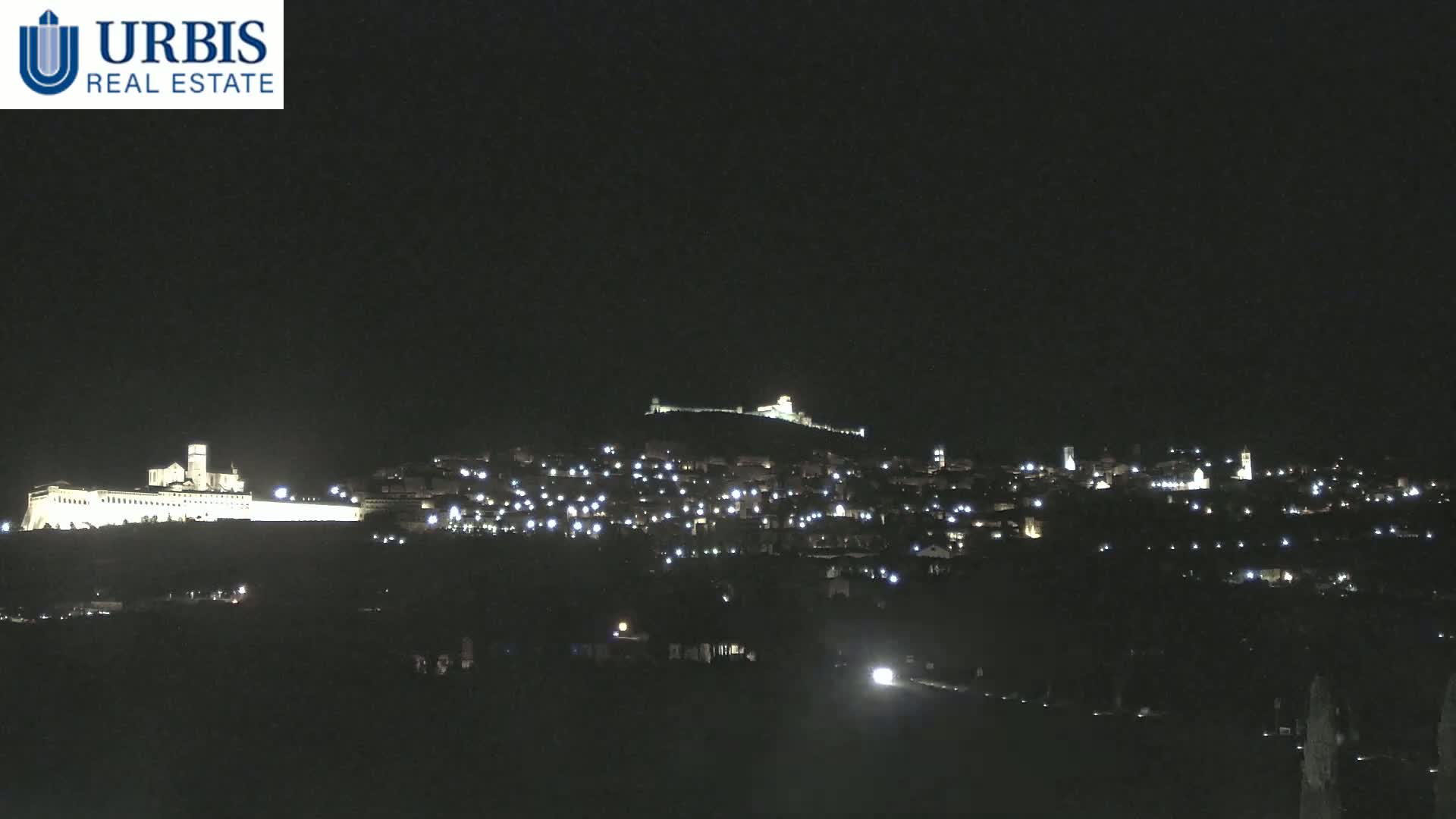 A nighttime view of a town illuminated by scattered lights on a hill, with a large, brightly lit building on a lower hill in the distance.