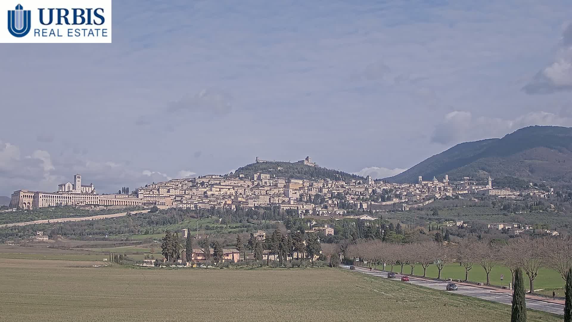 A large hilltop town, under a mostly clear sky, is viewed from a distance across a flat, sparsely vegetated field, with a road and several cars visible in the foreground.