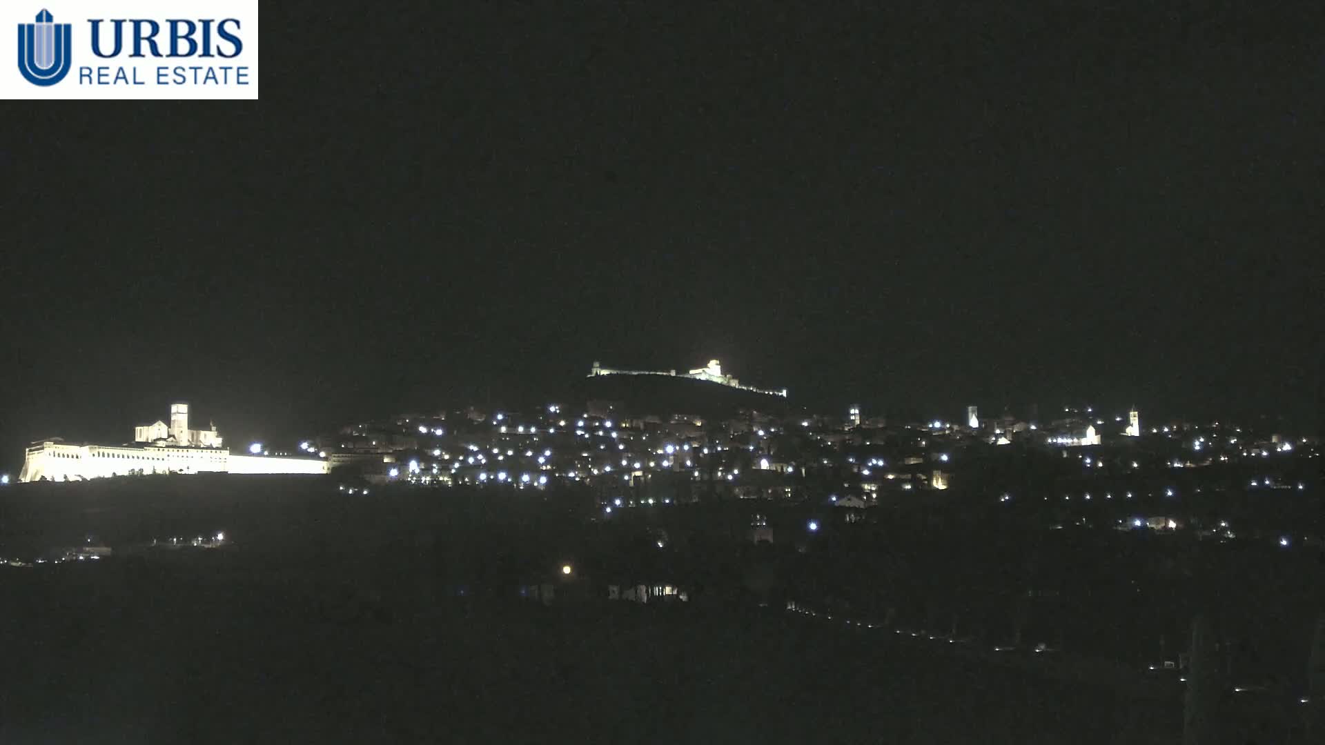 A nighttime view of a town nestled on a hill, illuminated by scattered lights, with a larger, brightly lit structure situated on a lower hill in the foreground.